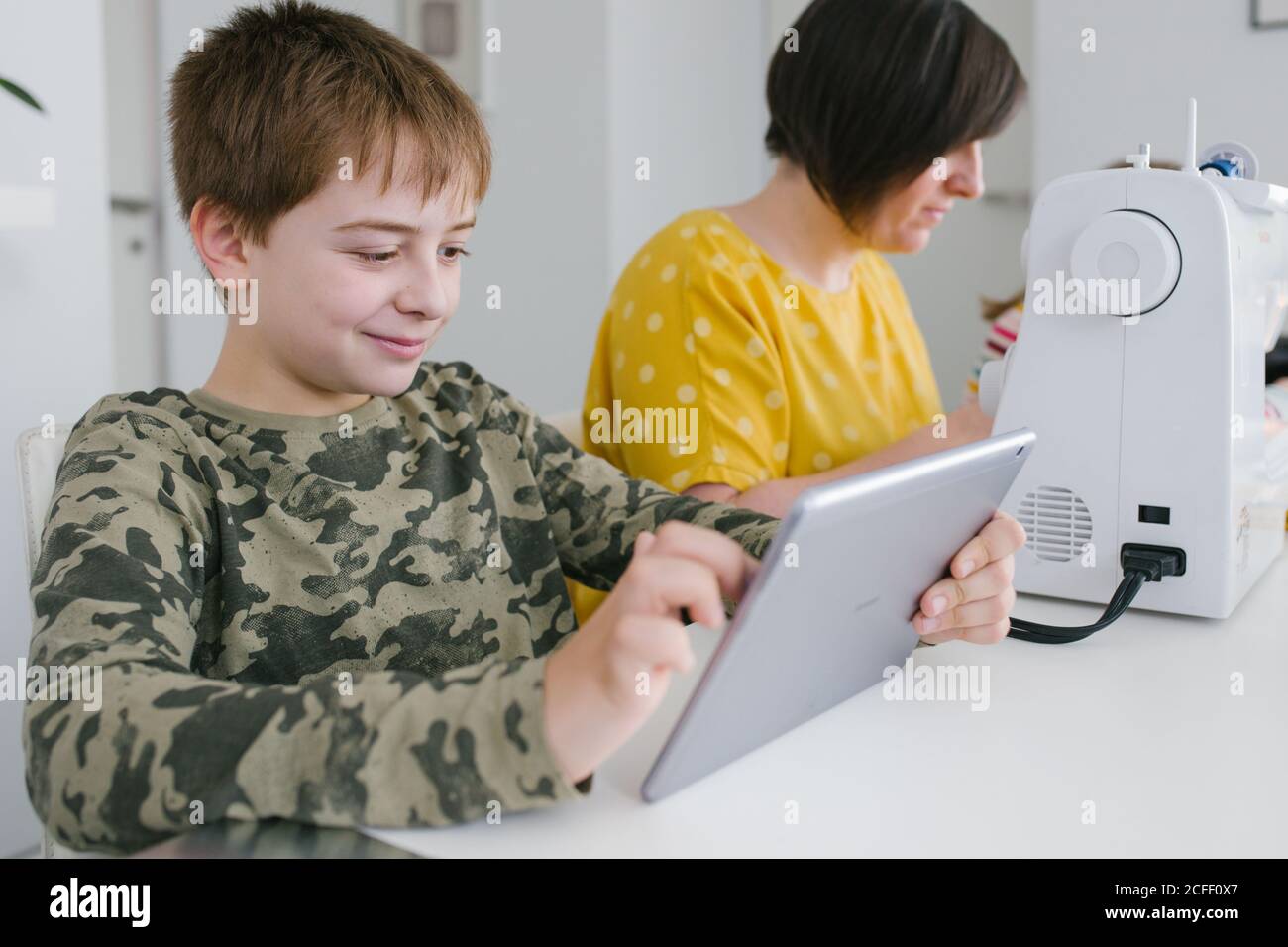 Adult Woman working in a sewing machine while little son browses on modern table sitting at table at home Stock Photo