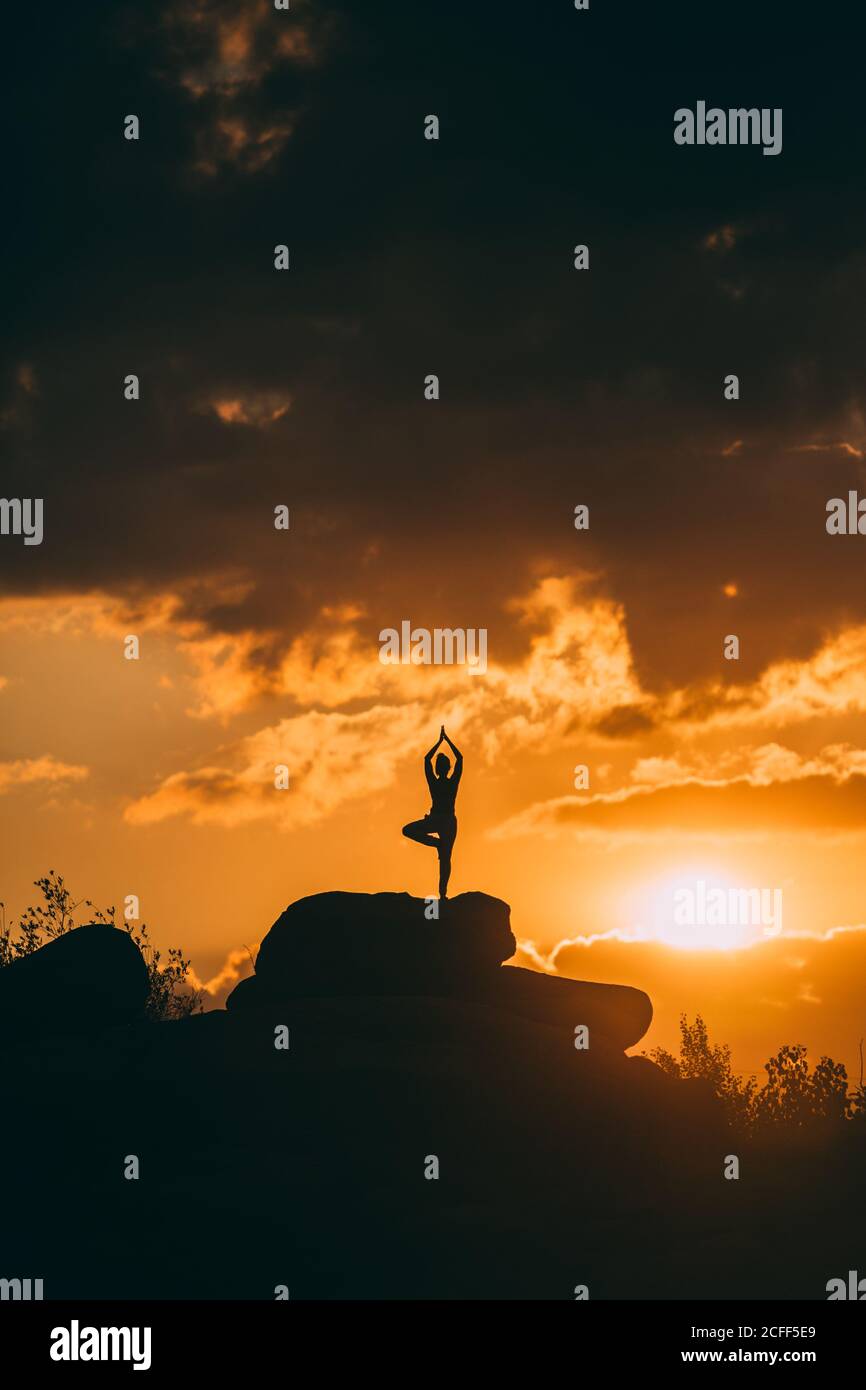 silhouette of Woman doing yoga poses outdoors on a amazing sunset while standing on rock Stock Photo