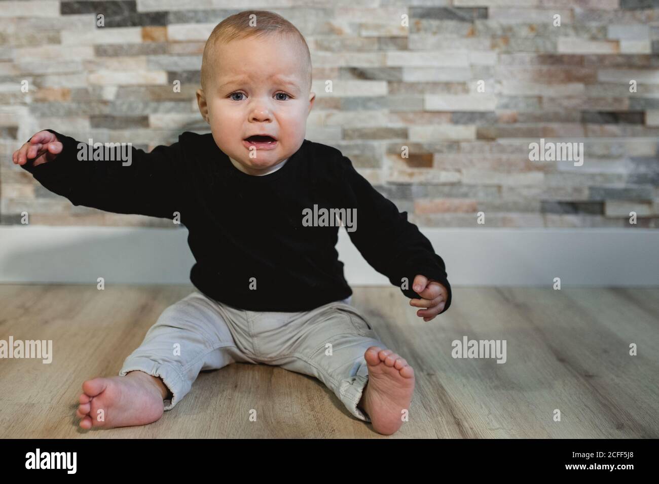 Upset barefoot baby in jumper and pants sitting on floor and crying against wall at home Stock Photo