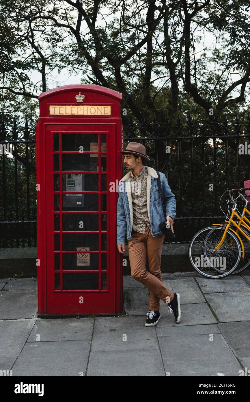 Relaxed male photographer in casual clothes and hat searching inspiration and view for shooting while leaning on traditional old style telephone call box against green trees on street in Scotland Stock Photo