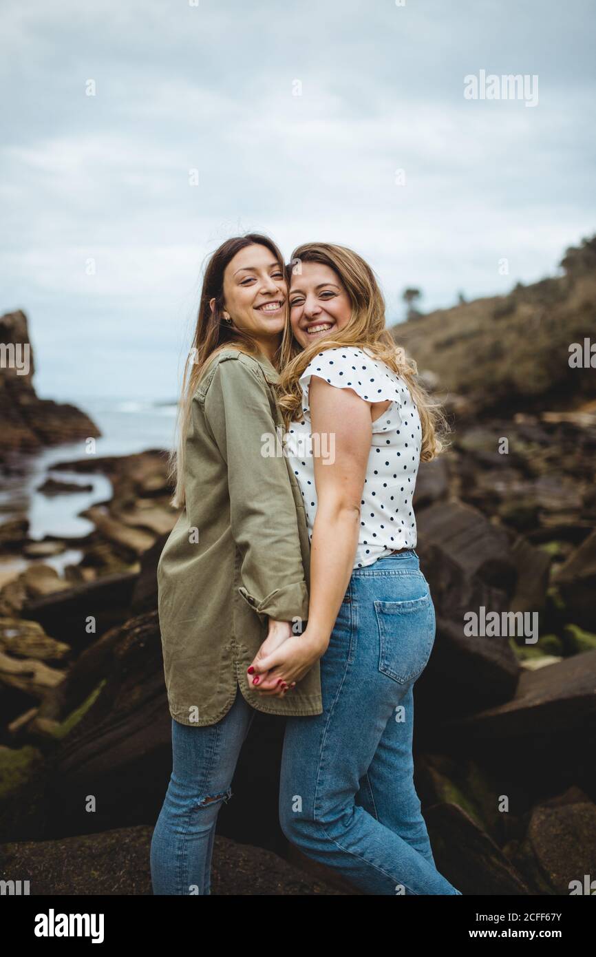 Relaxed women standing on stone on hill and hugging after walking in countryside on cloudy spring day and enjoying time together on cloudy spring day Stock Photo
