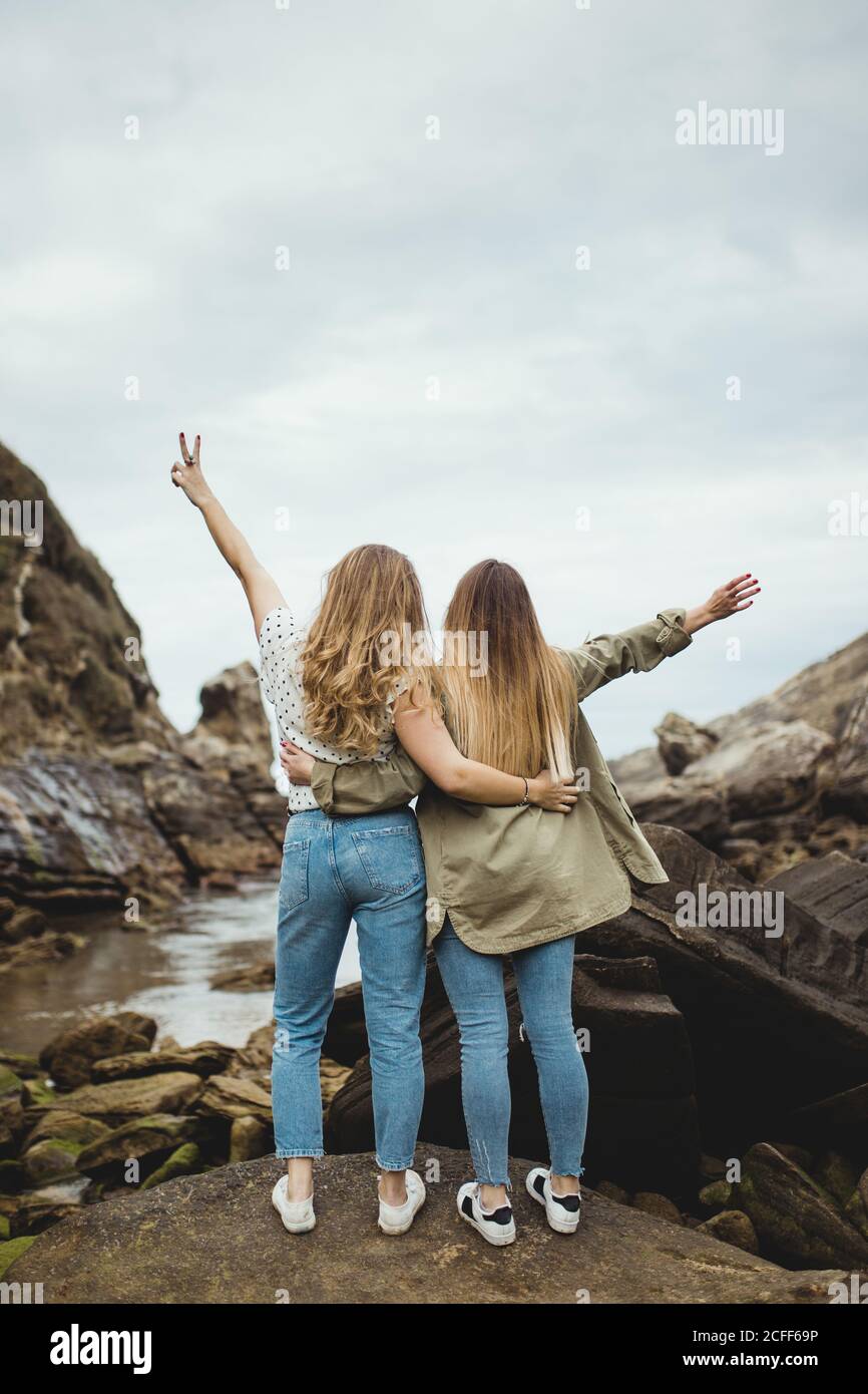 Back view of anonymous women standing on stone on hill and hugging after walking in countryside on cloudy spring day and enjoying time together on cloudy spring day Stock Photo