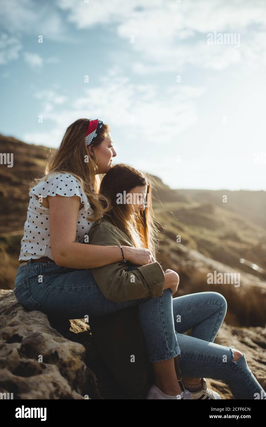 Relaxed women sitting on stone on hill and hugging while resting after walking in countryside on cloudy spring day and enjoying time together on cloudy spring day Stock Photo