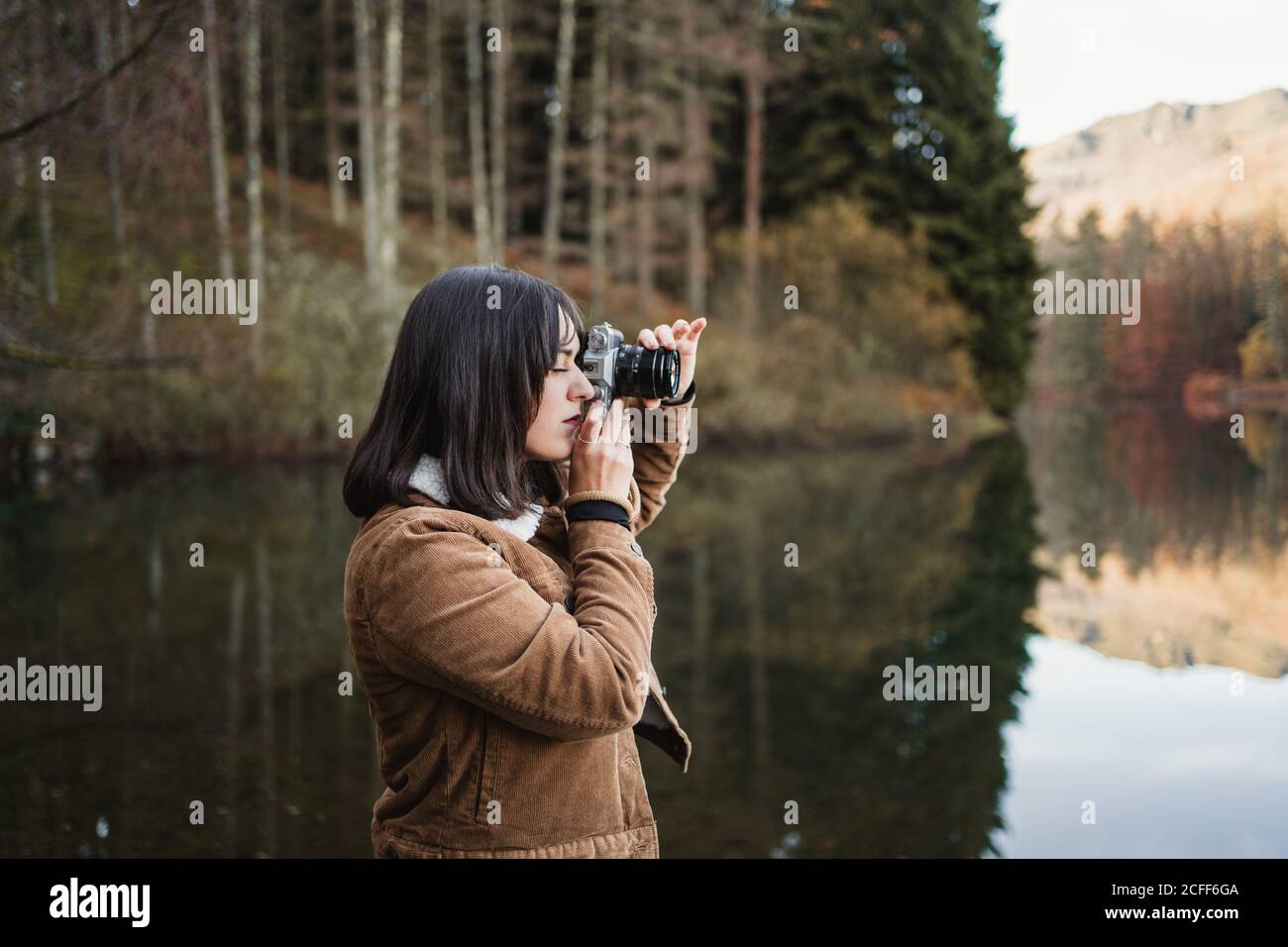 Side view of young female traveler in brown jacket taking picture with photo camera while standing next to lake surrounded with forest in autumn day Stock Photo