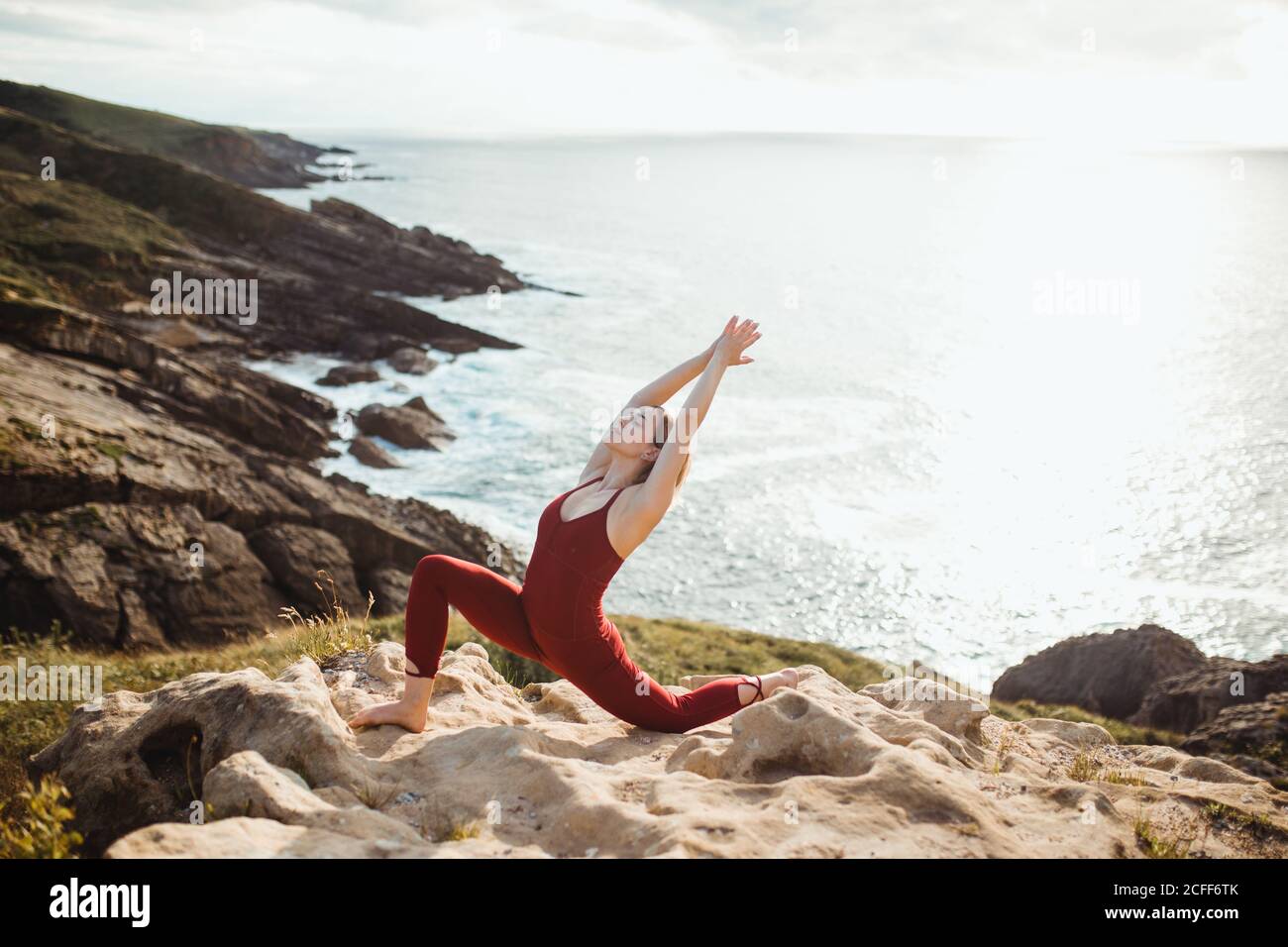 Side view of young blond haired Woman with closed eyes in red sporty wear working out and practicing warrior one yoga pose on empty rocky beach Stock Photo