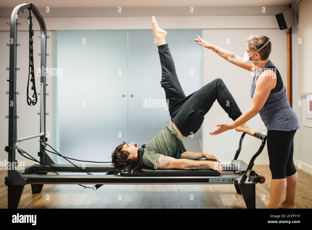 Concentrated male in sportswear doing exercises on pilates reformer during training under supervision of female instructor in protective respirator during coronavirus pandemic Stock Photo