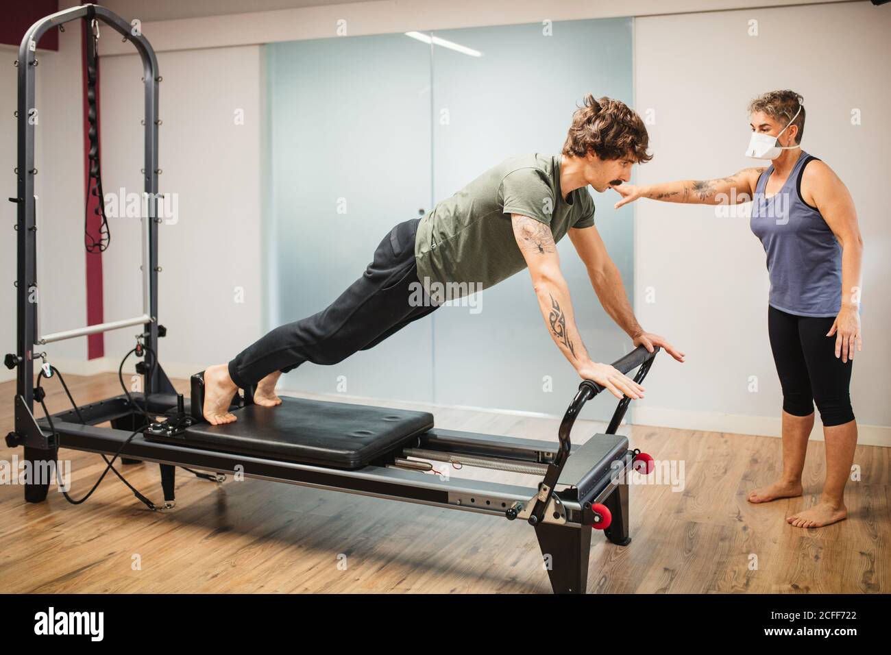 Concentrated male in sportswear doing exercises on pilates reformer during training under supervision of female instructor in protective respirator during coronavirus pandemic Stock Photo