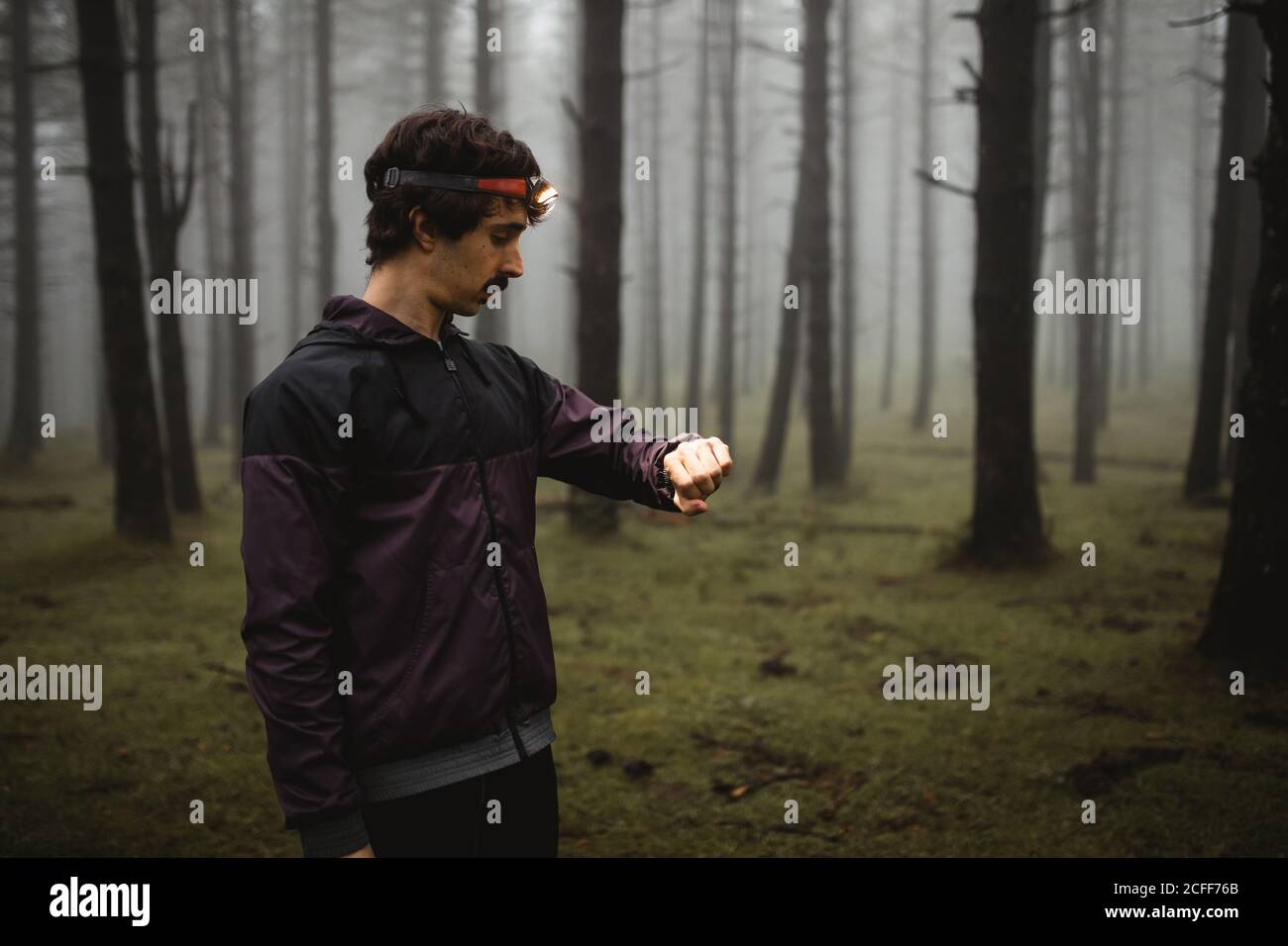Fit sportsman in activewear and head torch standing in foggy forest and checking time on wristwatch during morning workout Stock Photo