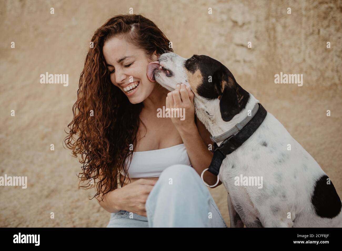 Funny dog licking cheek of excited young lady against weathered building wall on street Stock Photo