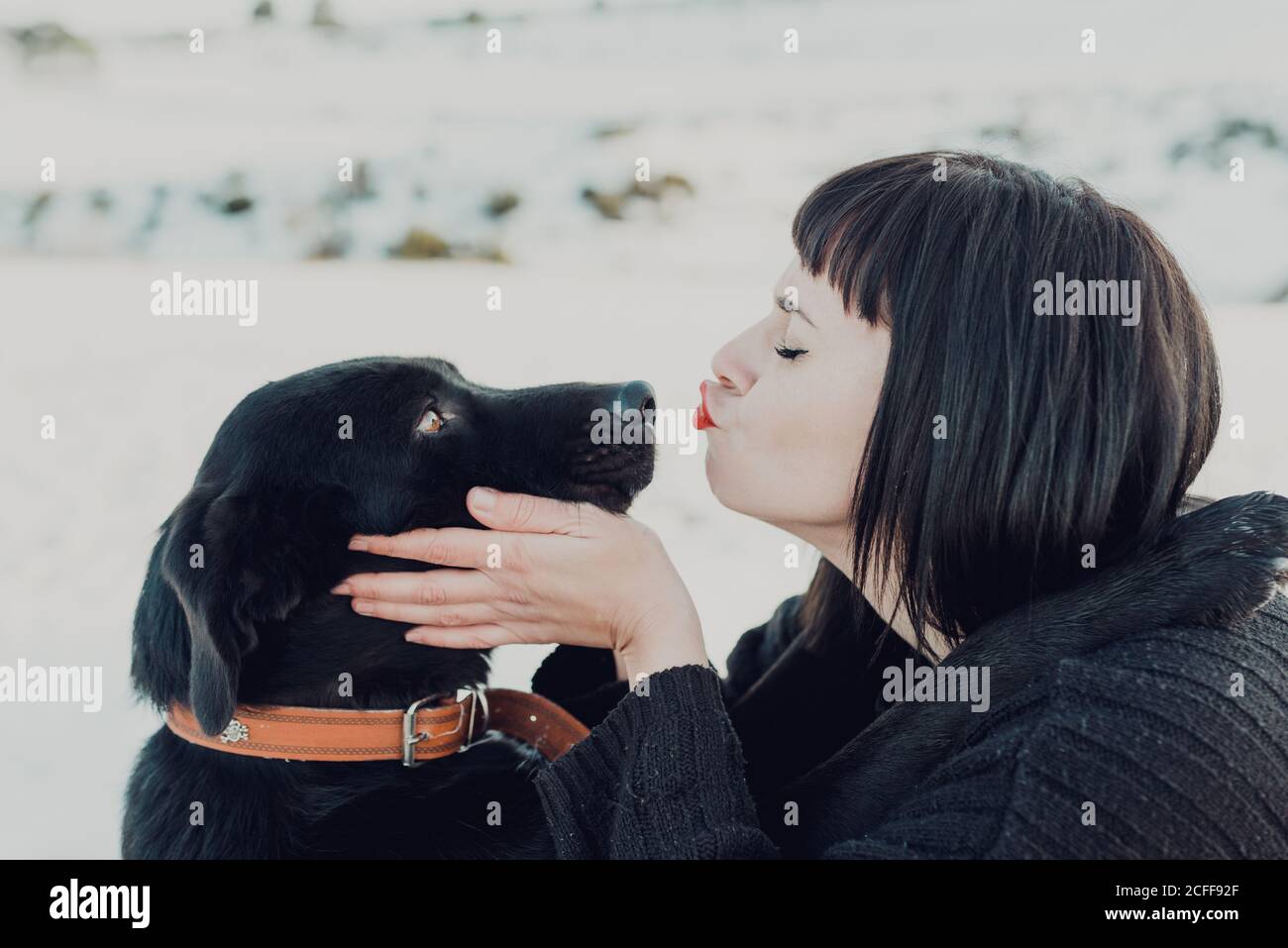 Side view of young brunette female with eyes closed kissing in nose adorable black dog Stock Photo