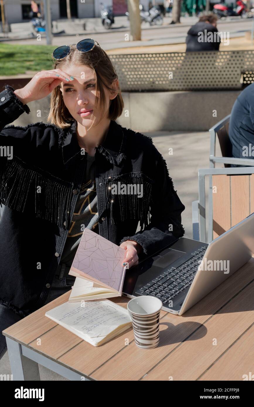 Young female freelancer sitting at table with open netbook and copybook during coffee break and looking away covering eyes from bright sunlight Stock Photo