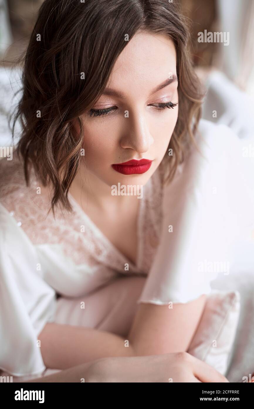Gorgeous Woman with red lips in white dress looking away while sitting on floor beside sofa Stock Photo