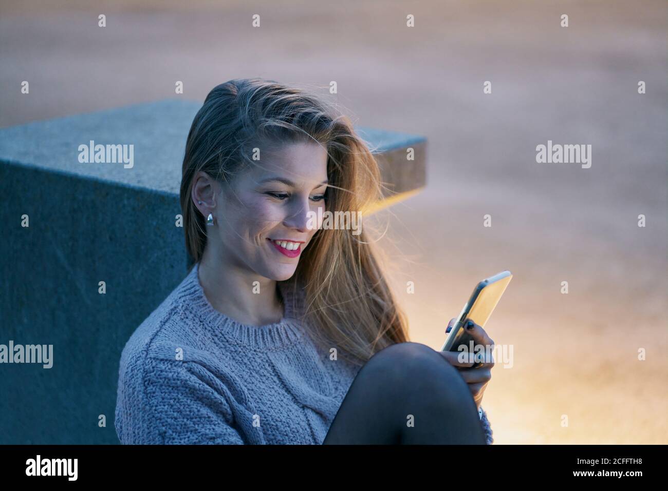 Happy young Woman in casual jumper watching social media on smartphone and smiling while sitting at stone bench in street Stock Photo