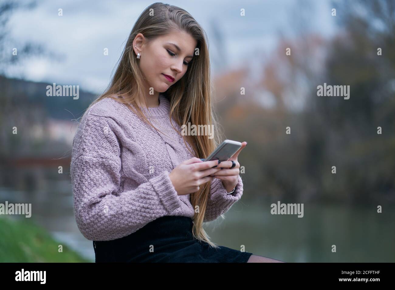 Happy young Woman in casual jumper watching social media on smartphone and smiling while sitting at stone bench in street Stock Photo