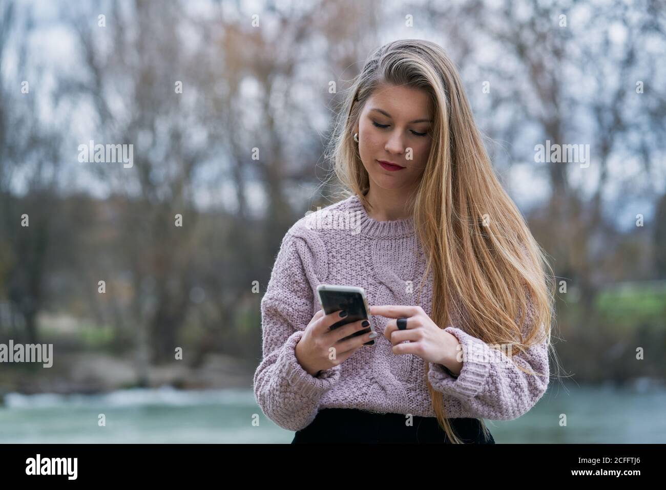Relaxed young Woman in casual jumper watching social media on smartphone and smiling while sitting at stone bench in street Stock Photo