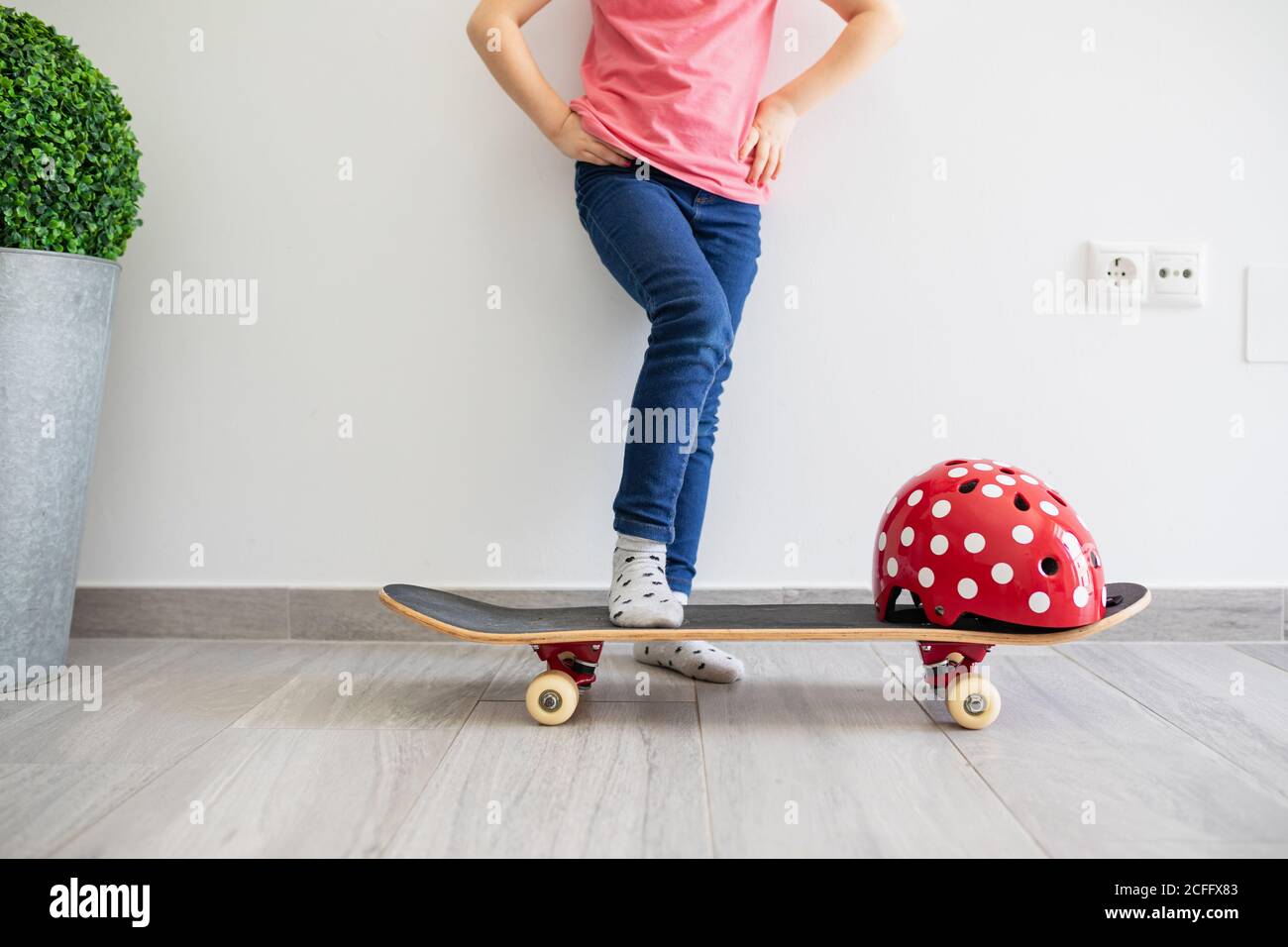 Low angle of faceless cool preteen kid in casual clothes with longboard and vivid red protective helmet in light contemporary flat dreaming about street staying at home Stock Photo