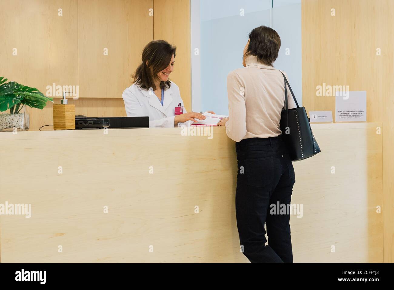 Smiling specialist in white uniform and with name tag reviewing documents while conversing with unrecognizable female patient at reception desk in clinic Stock Photo