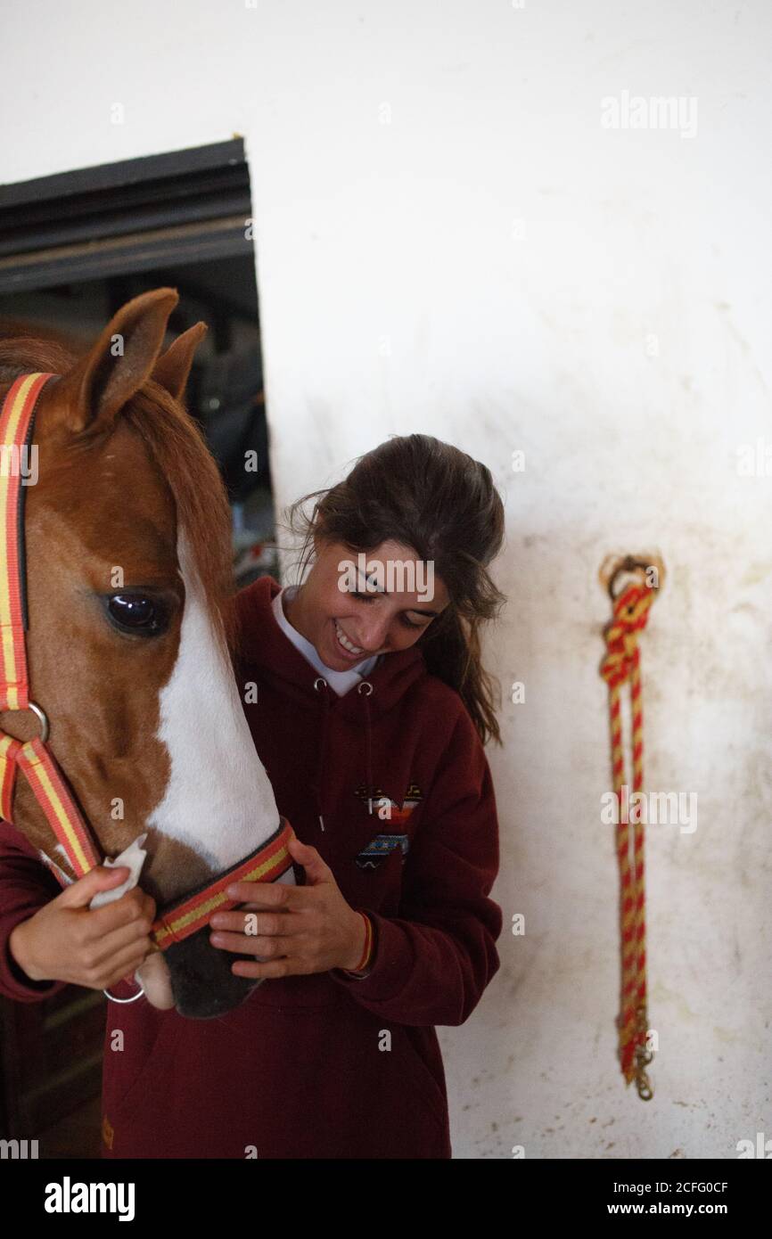 Teenage girl embracing with small pony in cute hat on ears standing inside of stable Stock Photo