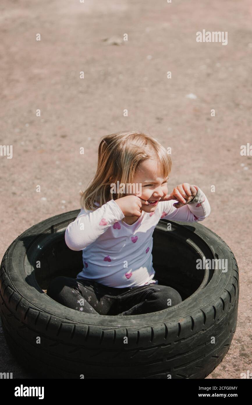 From above of happy adorable little girl sitting inside car tire while having fun and playing outdoors on summer day Stock Photo