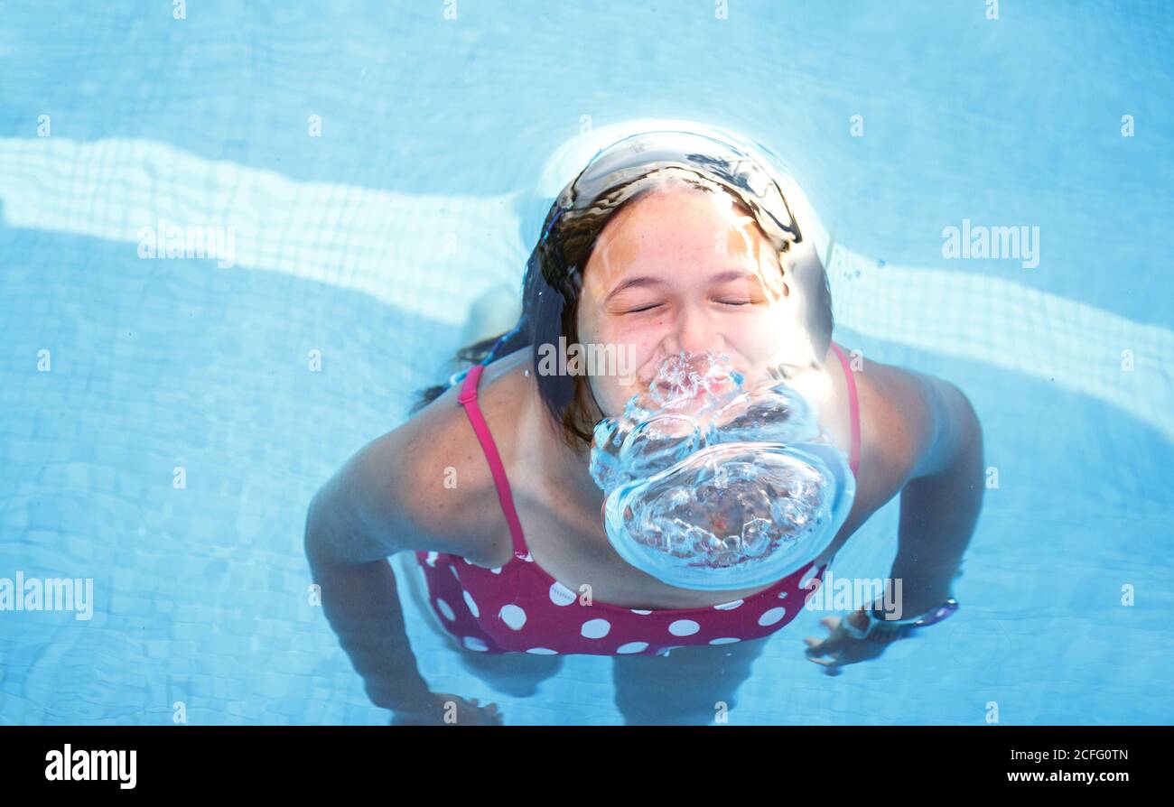From above cheerful teen girl blowing bubbles with nose under water while plunging in clean pool