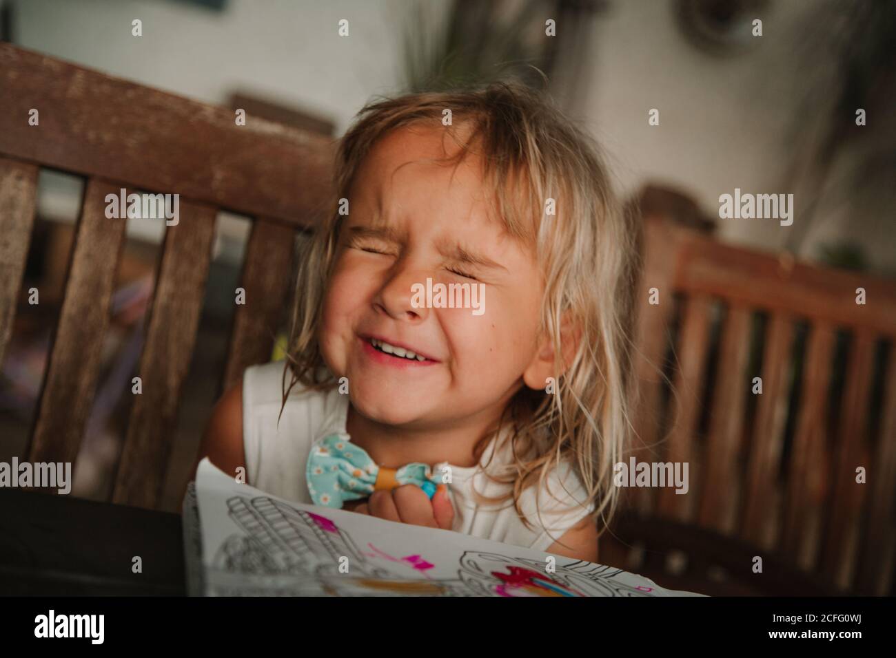 Adorable little girl squinting and smiling while sitting at table with coloring book and waiting for surprise at home Stock Photo