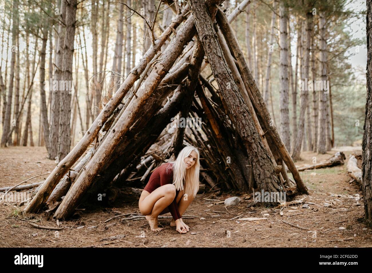 Side view of carefree blonde barefoot Woman in bright unitard sitting on hunkers next to hut in pine forest Stock Photo