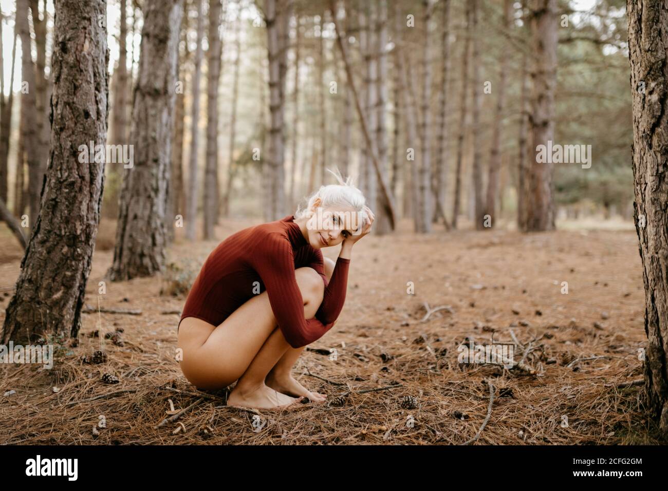 Side view of attractive blonde Woman in leotard squatting and embracing knees sitting barefoot in pine forest Stock Photo