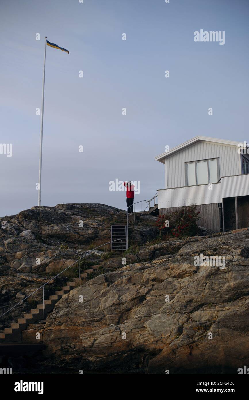 From below of anonymous male tourist in casual outfit taking photo of landscape while standing on stairway on mount near house in autumn Stock Photo