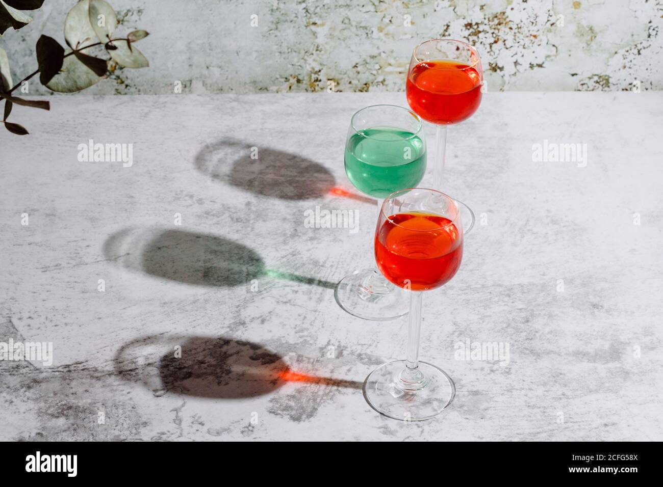 From above of glasses filled with colorful liquids put on concrete surface near plant with dark green leaves and rusty wall Stock Photo