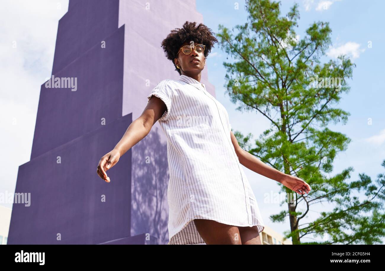 From below pensive relaxed African American female in glasses and white dress looking at camera while standing alone against purple facade of high rise building and evergreen tree in sunny day Stock Photo