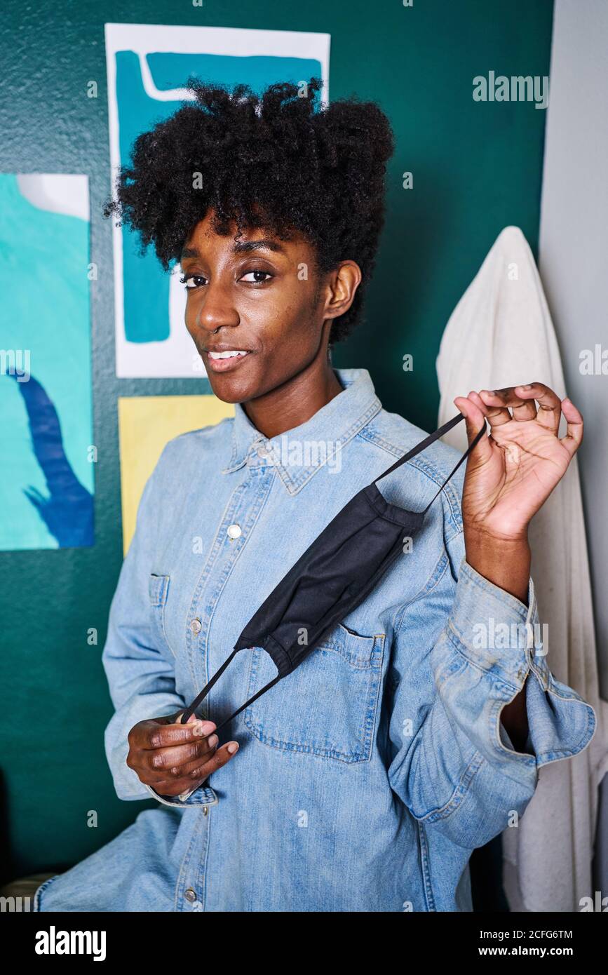 Positive young African American female in casual wear looking at camera demonstrating black protective mask for coronavirus prevention while standing near wall in modern room at home Stock Photo