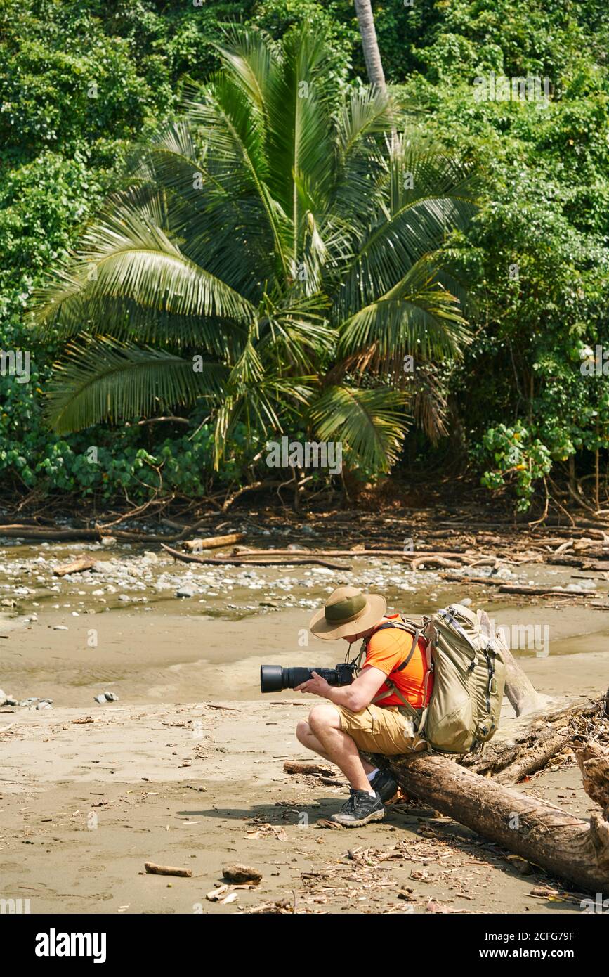 Side view of anonymous male tourist with backpack taking photos of landscape with professional camera while sitting in a tree branch on shore in Costa Rica Stock Photo