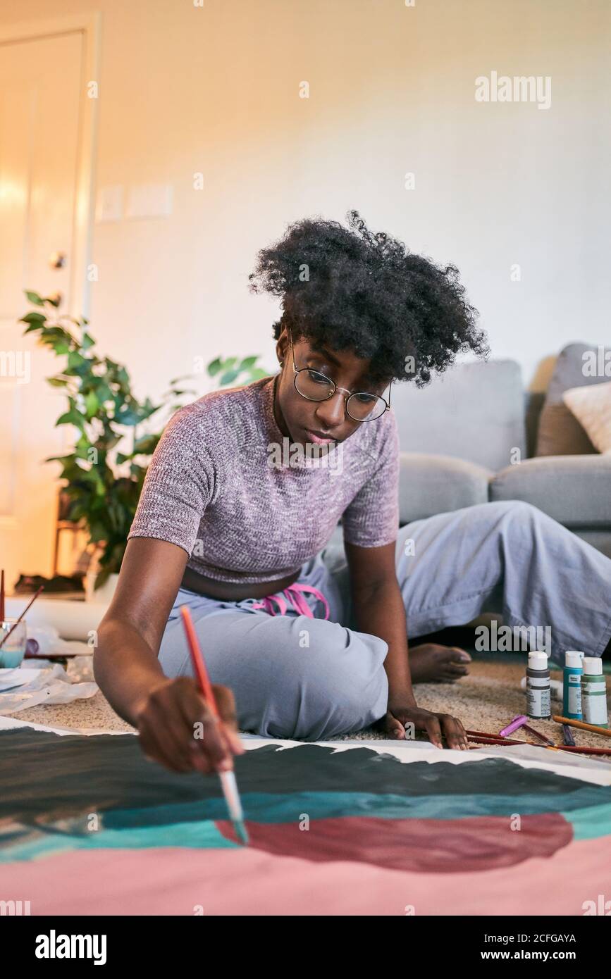 Young beautiful African American Woman with afro hair, amateur artist in casual outfit drawing colorful picture with paintbrush on paper while sitting on floor in cozy apartment Stock Photo