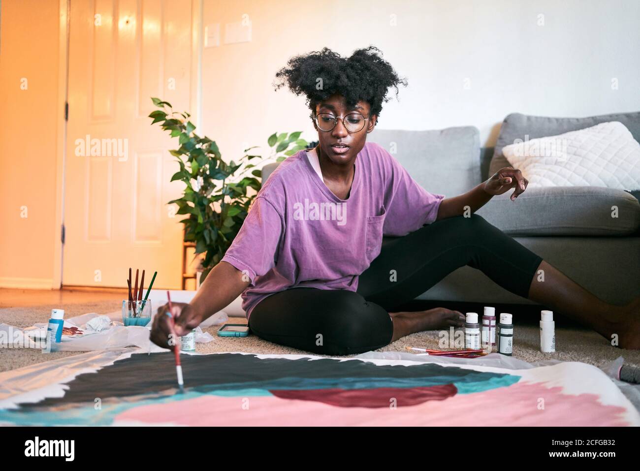 Young happy beautiful African American Woman, amateur artist in casual outfit drawing colorful picture with paintbrush on paper while sitting on floor in cozy apartment Stock Photo