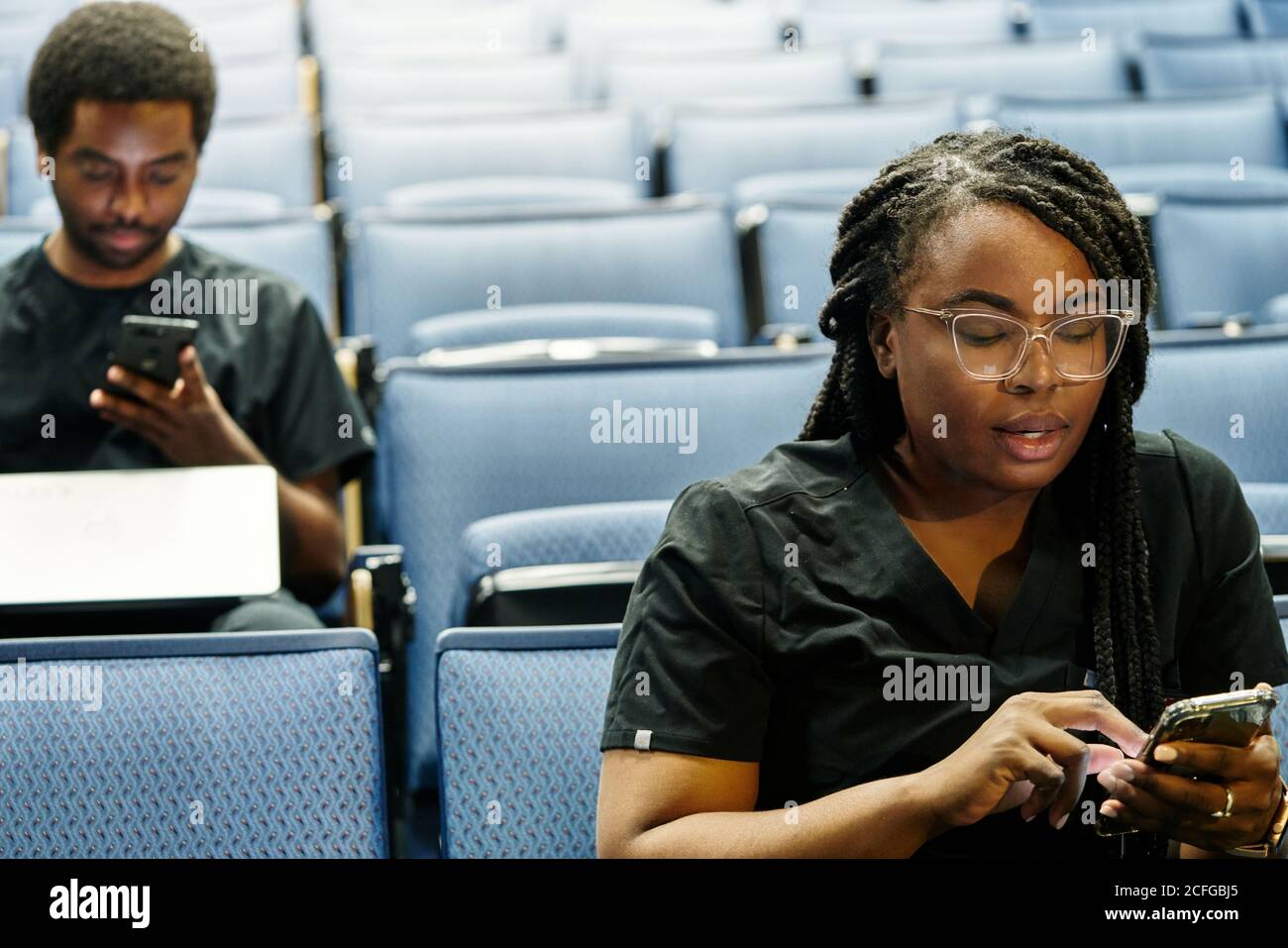 Black woman with braids and African American man sitting in auditorium and browsing smartphones during lesson in auditorium Stock Photo