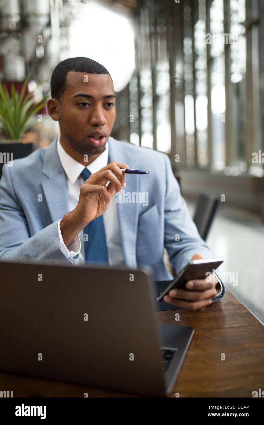 Concentrated black man calculating in mind sitting at desk with laptop and smartphone looking away. Stock Photo