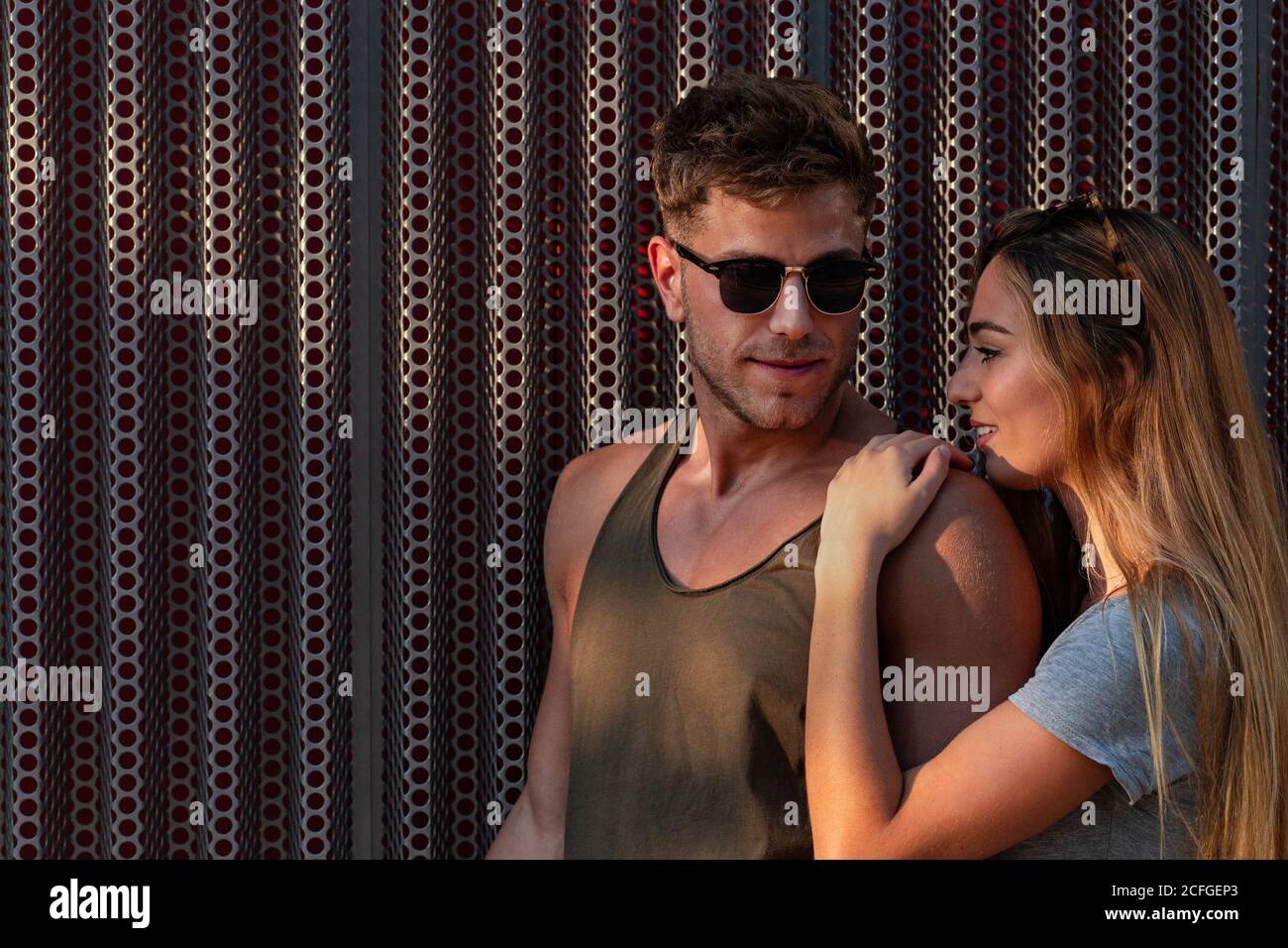 Handsome young man and pretty Woman smiling and looking at each other while standing near metal wall on street and embracing Stock Photo