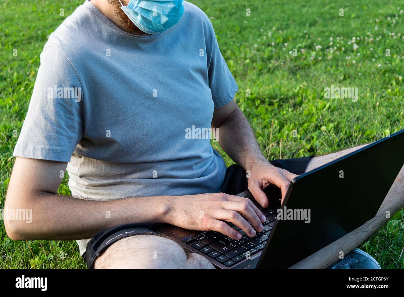 A caucasian bearded man is sitting on the grass in the sun with his laptop. Wearing surgical mask, prevent coronavirus, smart working on nature Stock Photo