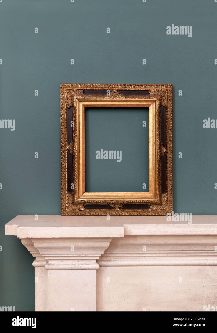 Gilded or golden empty rectangular vintage frame on a mantelpiece for a picture or painting leaning against a green wall Stock Photo