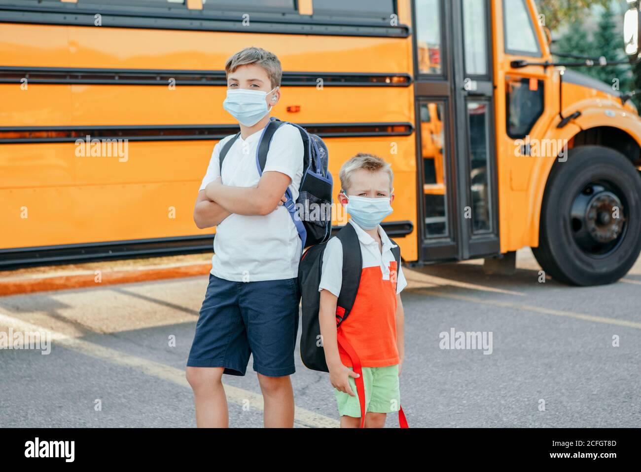 Sad brothers students wearing face mask near yellow bus. Kids with personal protective equipment on face. Education and back to school in September. Stock Photo