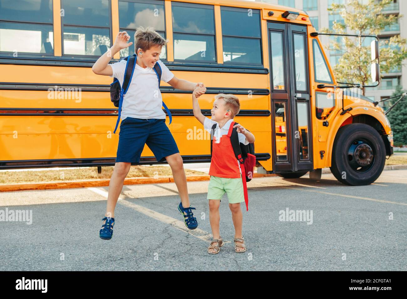 Happy Caucasian brothers students jumping near yellow school bus. Smiling kids going back to school in September. Education system and learning. Suppo Stock Photo