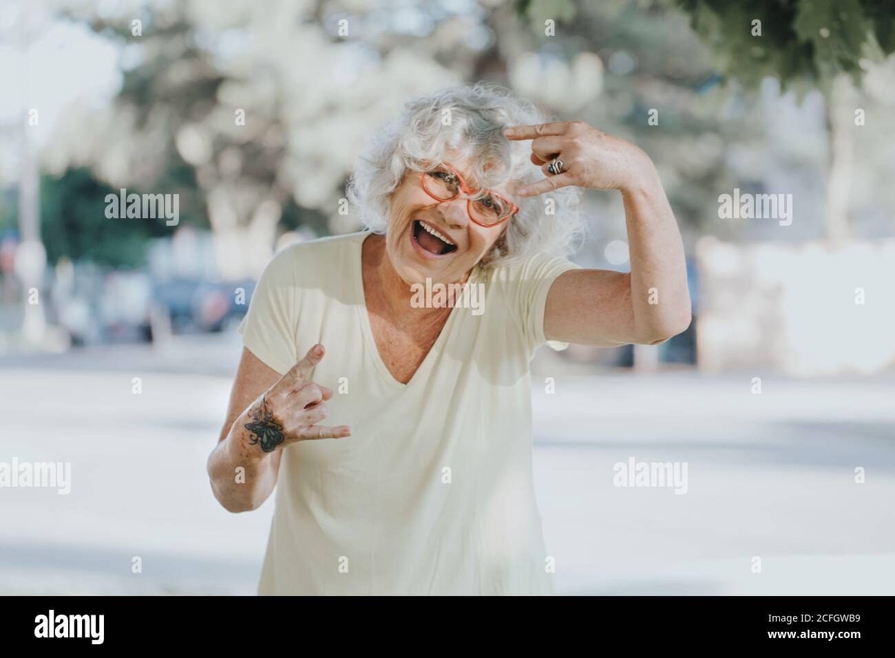 Cool edgy Caucasian old lady woman with butterfly tattoo on hand. Beautiful funky elderly woman with white grey hair making devil rock sign Stock Photo