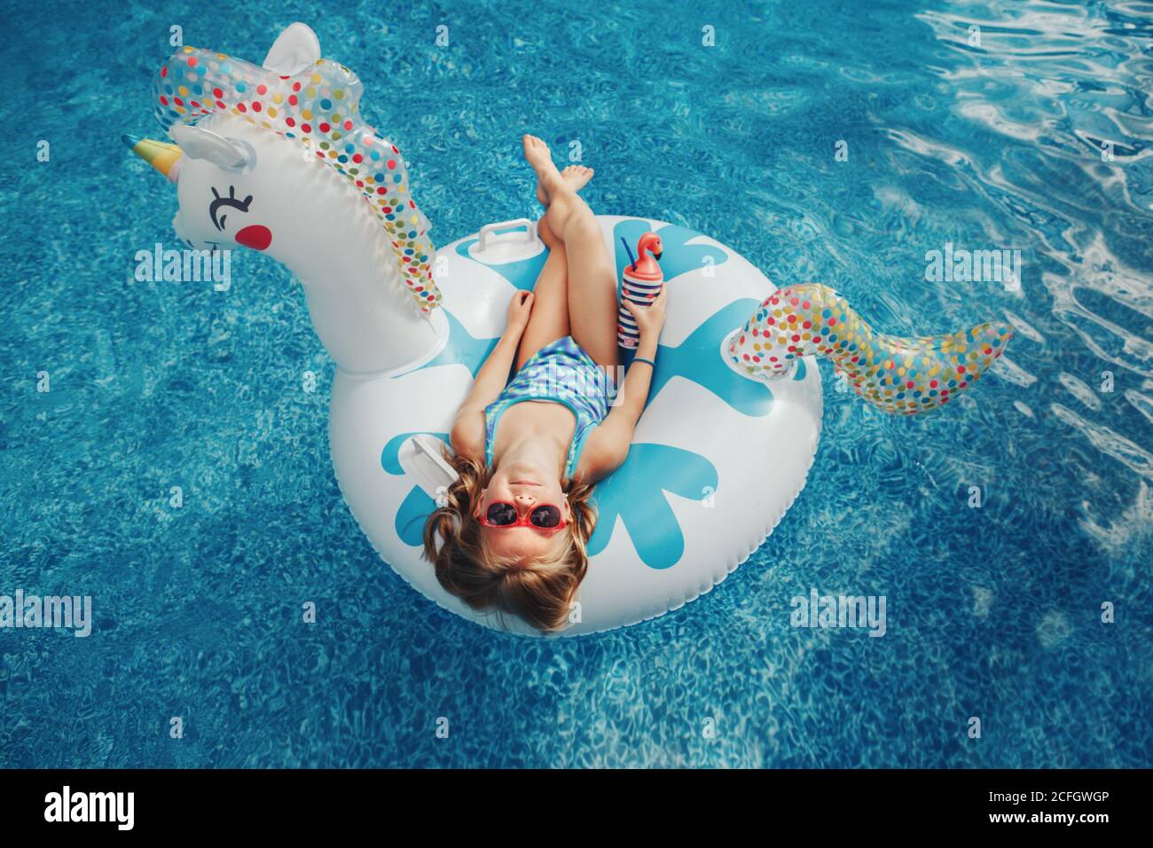 Cute adorable girl in sunglasses with drink lying on inflatable ring unicorn. Kid child enjoying having fun in swimming pool. Summer outdoor water act Stock Photo