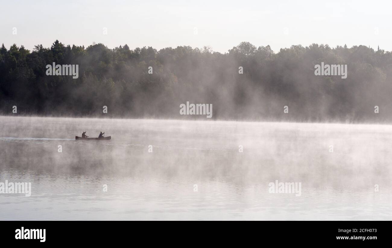 Misty Morning Paddle: Two people paddle a canoe through the early morning mist on a calm lake as the rising sun highlights the rising fog from the warm lake. Stock Photo