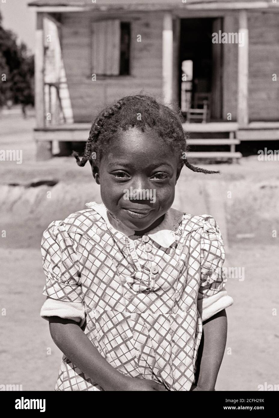 1930s SHY SMILING AFRICAN-AMERICAN LITTLE GIRL LOOKING AT CAMERA STANDING IN FRONT OF ROUGH RURAL CABIN HOME SOUTH CAROLINA USA - n1736 HAR001 HARS HOME LIFE UNITED STATES COPY SPACE HALF-LENGTH INSPIRATION UNITED STATES OF AMERICA CABIN ROUGH B&W NORTH AMERICA EYE CONTACT NORTH AMERICAN HAPPINESS WELLNESS CHEERFUL AFRICAN-AMERICANS AFRICAN-AMERICAN BLACK ETHNICITY PRIDE IN OF SMILES CONCEPTUAL JOYFUL SHY STYLISH DISADVANTAGED GROWTH JUVENILES SC SOUTH CAROLINA BLACK AND WHITE ERA HAR001 OLD FASHIONED AFRICAN AMERICANS Stock Photo