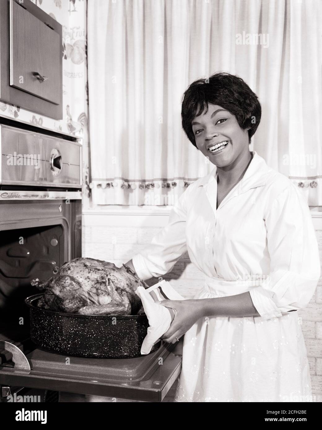 1960s SMILING AFRICAN-AMERICAN HOUSEWIFE IN KITCHEN LOOKING AT CAMERA TAKING A ROASTED TURKEY OUT OF A WALL OVEN  - n1859 HAR001 HARS HOME LIFE COPY SPACE HALF-LENGTH LADIES PERSONS CONFIDENCE AMERICANA B&W EYE CONTACT GOALS HOMEMAKER HAPPINESS HOMEMAKERS CHEERFUL AFRICAN-AMERICANS AFRICAN-AMERICAN EXCITEMENT BLACK ETHNICITY PRIDE ROASTED HOUSEWIVES SMILES THANKFUL CONNECTION THURSDAY JOYFUL NATIONAL HOLIDAY STYLISH GRATEFUL MID-ADULT MID-ADULT WOMAN NOVEMBER BLACK AND WHITE FOOD PREPARATION HAR001 OLD FASHIONED AFRICAN AMERICANS Stock Photo