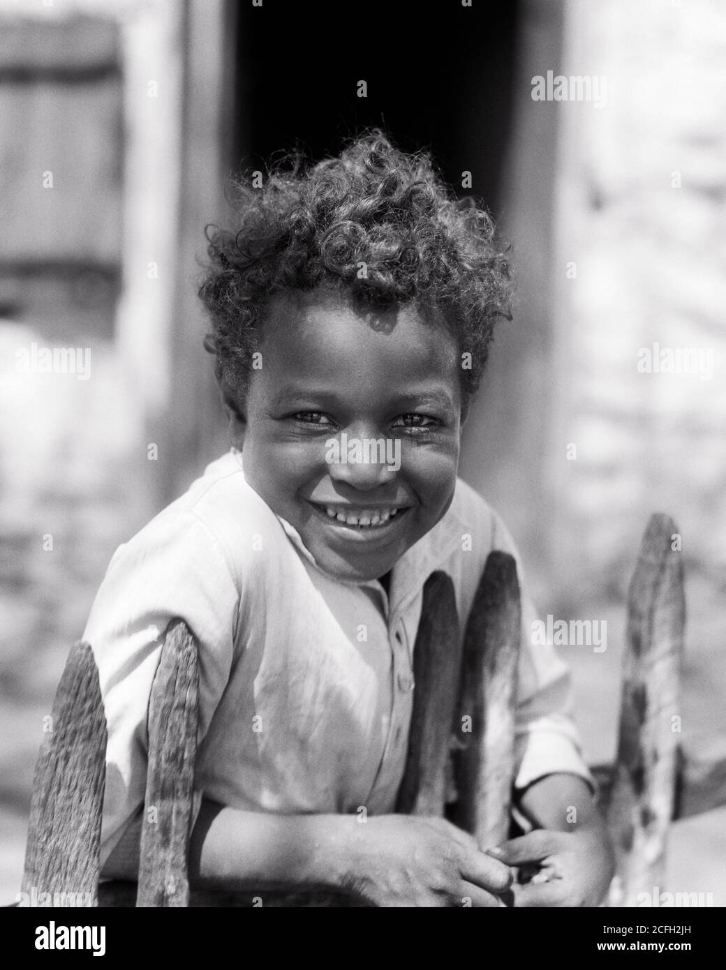 1930s SMILING DEPRESSION ERA CURLY HAIR AFRICAN-AMERICAN BOY LOOKING AT CAMERA LEANING ON WOOD PICKET FENCE IN FRONT OF HOME - n966 HAR001 HARS LIFESTYLE POOR RURAL HOME LIFE COPY SPACE HALF-LENGTH MALES CONFIDENCE EXPRESSIONS B&W EYE CONTACT HAPPINESS CHEERFUL STRENGTH AFRICAN-AMERICANS AFRICAN-AMERICAN BLACK ETHNICITY CURLY PRIDE IN OF ON SMILES CONCEPTUAL JOYFUL DISADVANTAGED GROWTH JUVENILES BLACK AND WHITE ERA HAR001 OLD FASHIONED AFRICAN AMERICANS Stock Photo