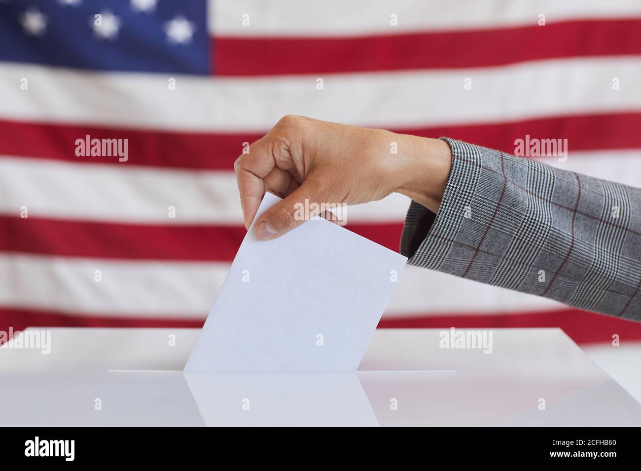 Side view close up of unrecognizable woman putting vote bulletin in ballot box while standing against American flag on election day, copy space Stock Photo