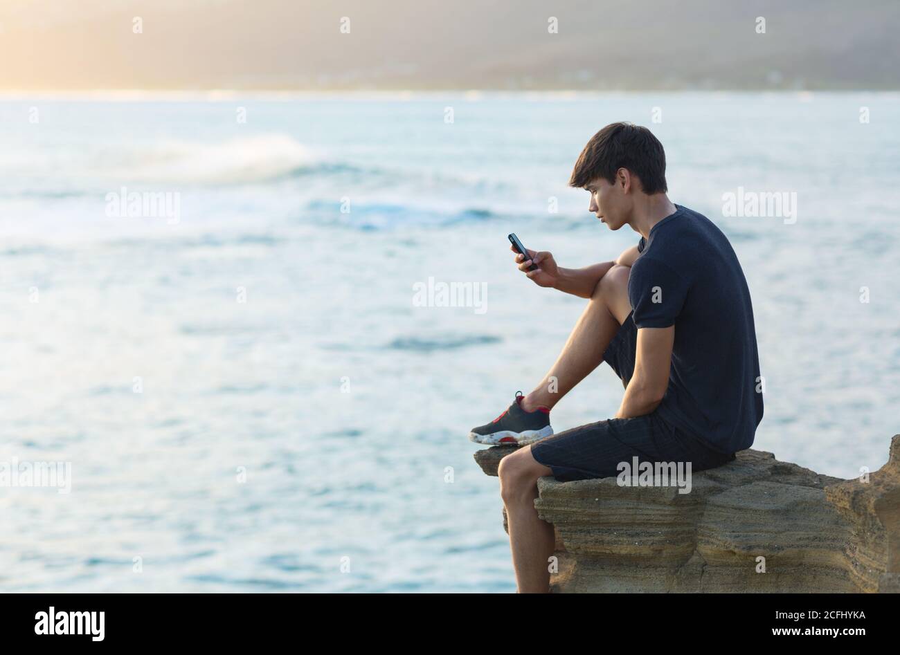 Teen boy using a mobile phone sitting on the edge on top of a cliff with the ocean view in the background during sunset. Communication and youth. Stock Photo