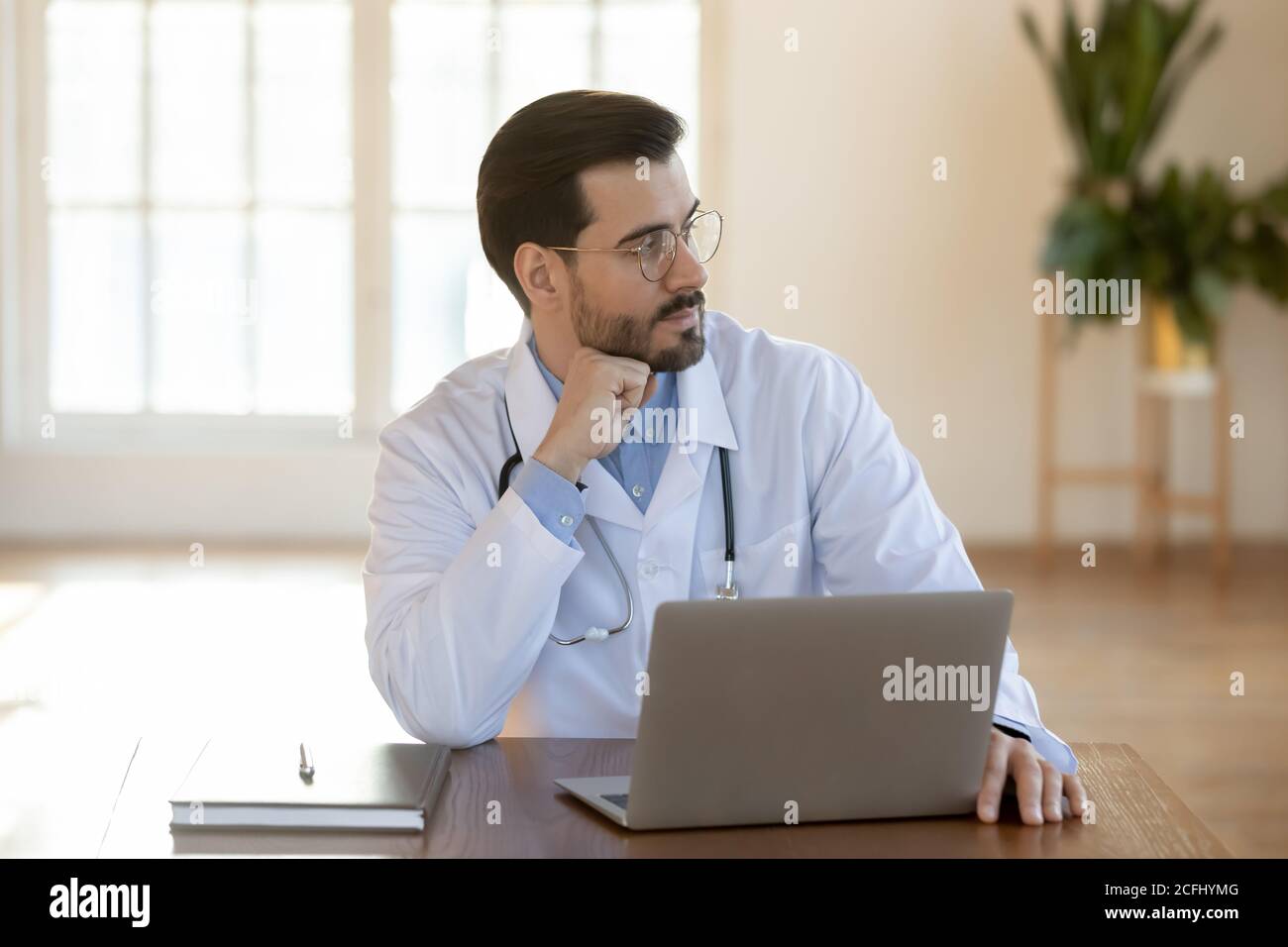 Thoughtful doctor wearing glasses working on laptop, pondering problem solution Stock Photo